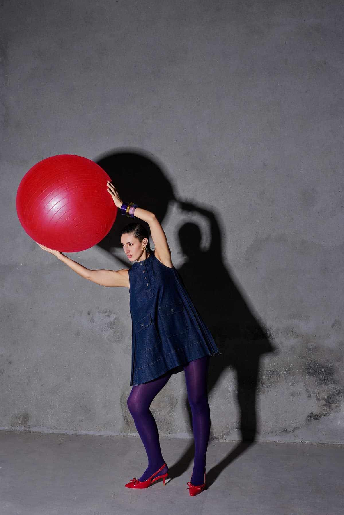 Model in a denim dress holding a red exercise ball against a concrete wall